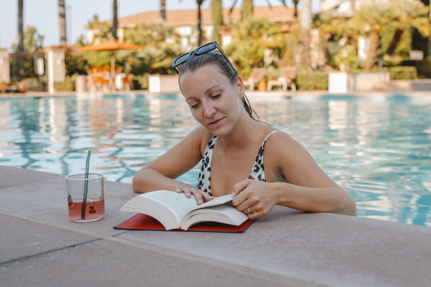 Someone enjoys reading a book and sipping a cocktail by a resort swimming pool on a sunny day in Huntington Beach.