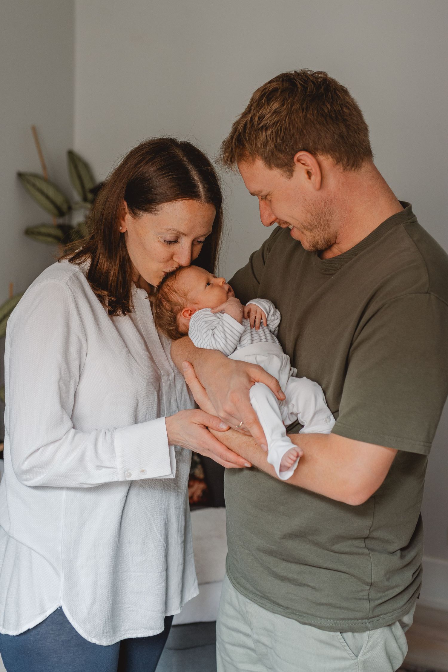 Parents sharing tender moments with their newborn baby against a neutral wall in candid lifestyle portraits at home in Huntington Beach.