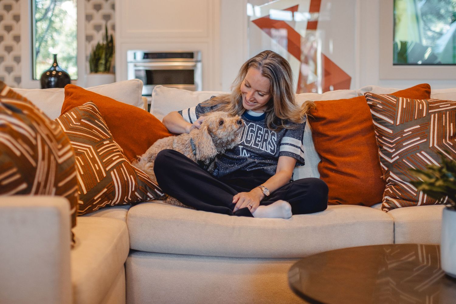 A cozy scene on a beige couch with orange pillows shows a person relaxing with a small dog in a modern living room in Huntington Beach.