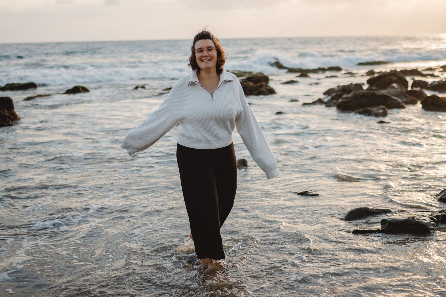 A person walks thoughtfully along a rocky ocean shoreline at sunset, waves crashing against the rocks in Orange County.