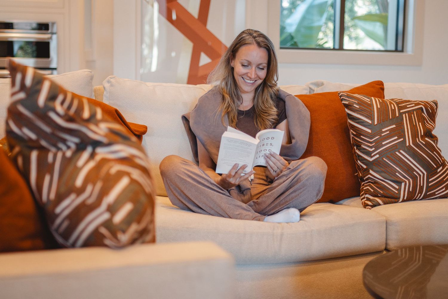 A person lounges comfortably on a brown sofa while reading a book in a sunlit living room in Huntington Beach.