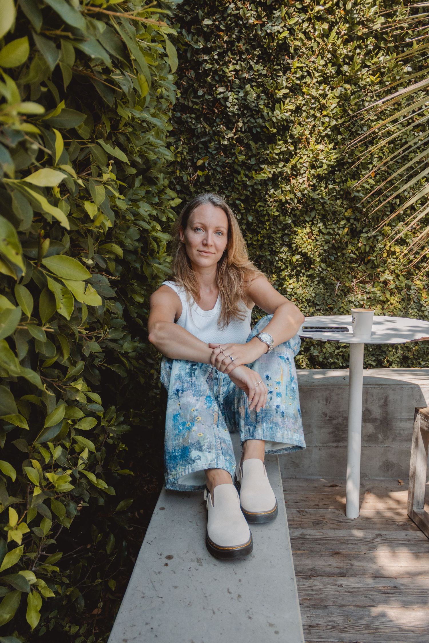 Person in white top and blue patterned skirt sits on concrete ledge in garden setting with white shoes in Laguna Beach.