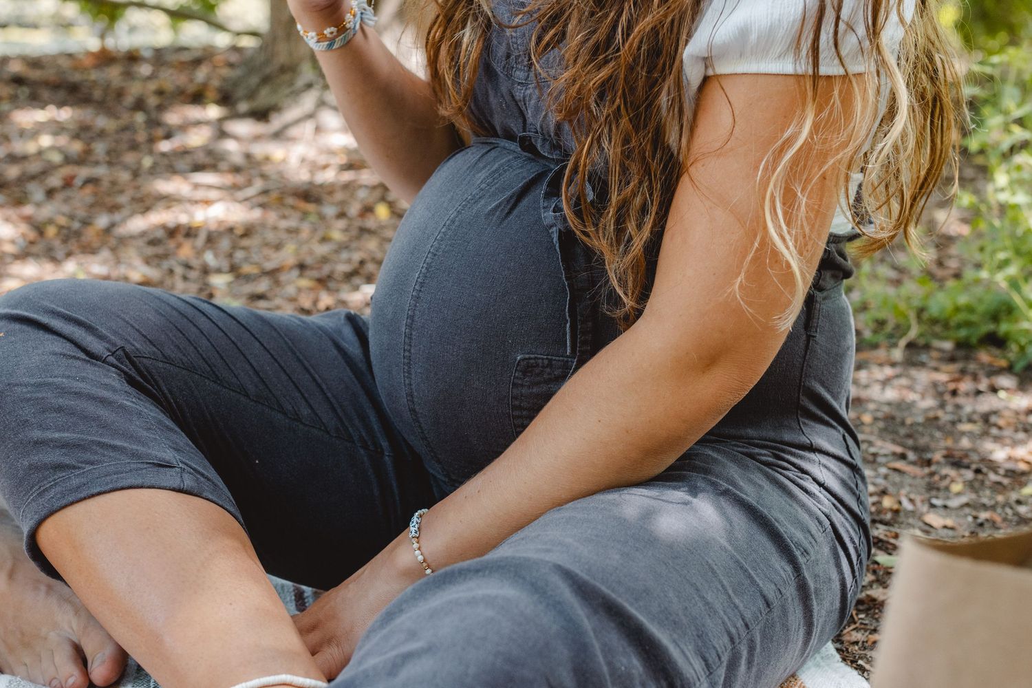 Pregnant women in casual attire relax on a fallen log in Newport Beach in a sunny wooded area outdoors.