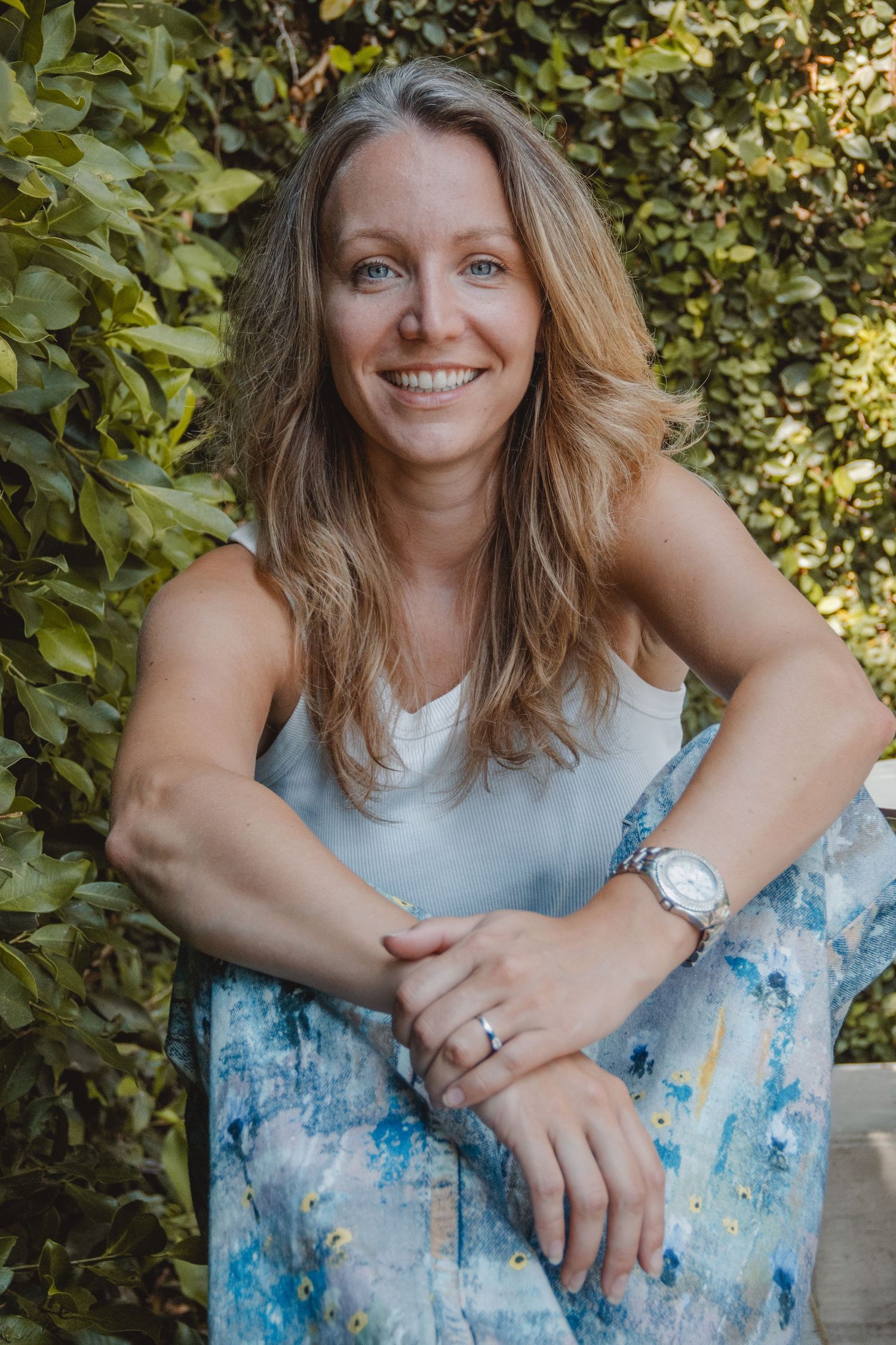 A person wearing a white top and blue patterned dress poses against a leafy green background in natural sunlight in Costa Mesa.