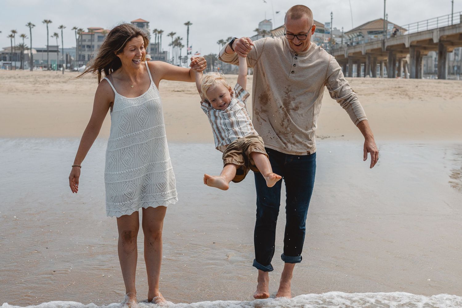 Couple with their kid walks along beach sand and water during golden hour creating candid romantic moment at sunset in Huntington Beach.