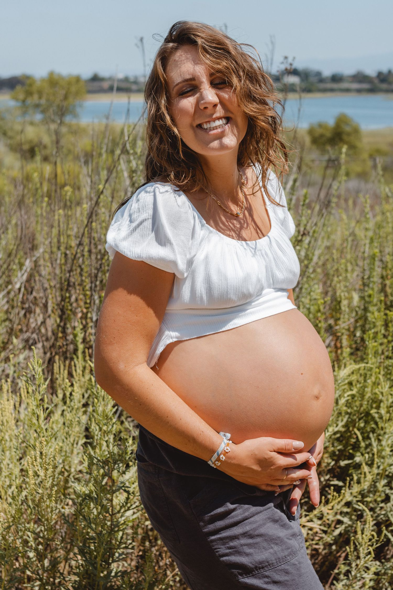 A smiling expectant mother wearing a white blouse and dark skirt poses in a field of tall grass in Newport Beach against a bright blue sky.