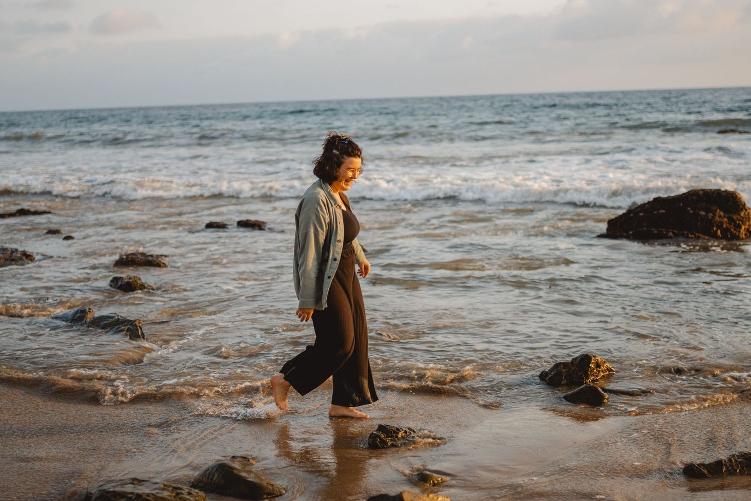 A person in a gray-green blazer walks along a rocky beach shoreline during golden hour in Laguna Beach.