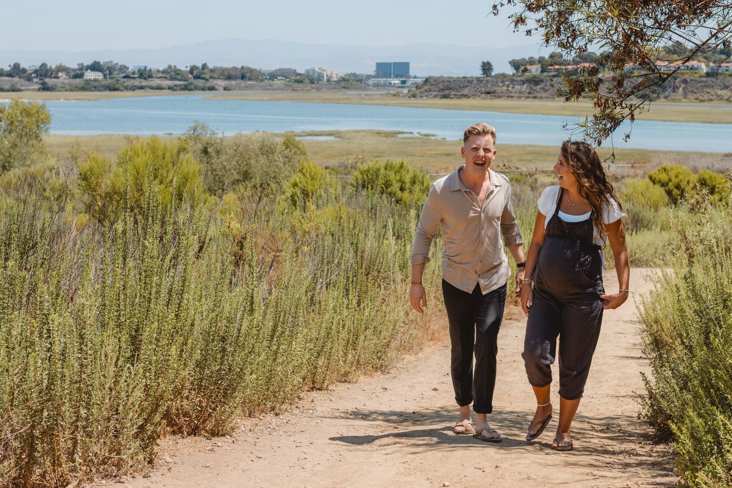 People walking along sandy trail through coastal wetlands in Newport Beach during a calm summer day.