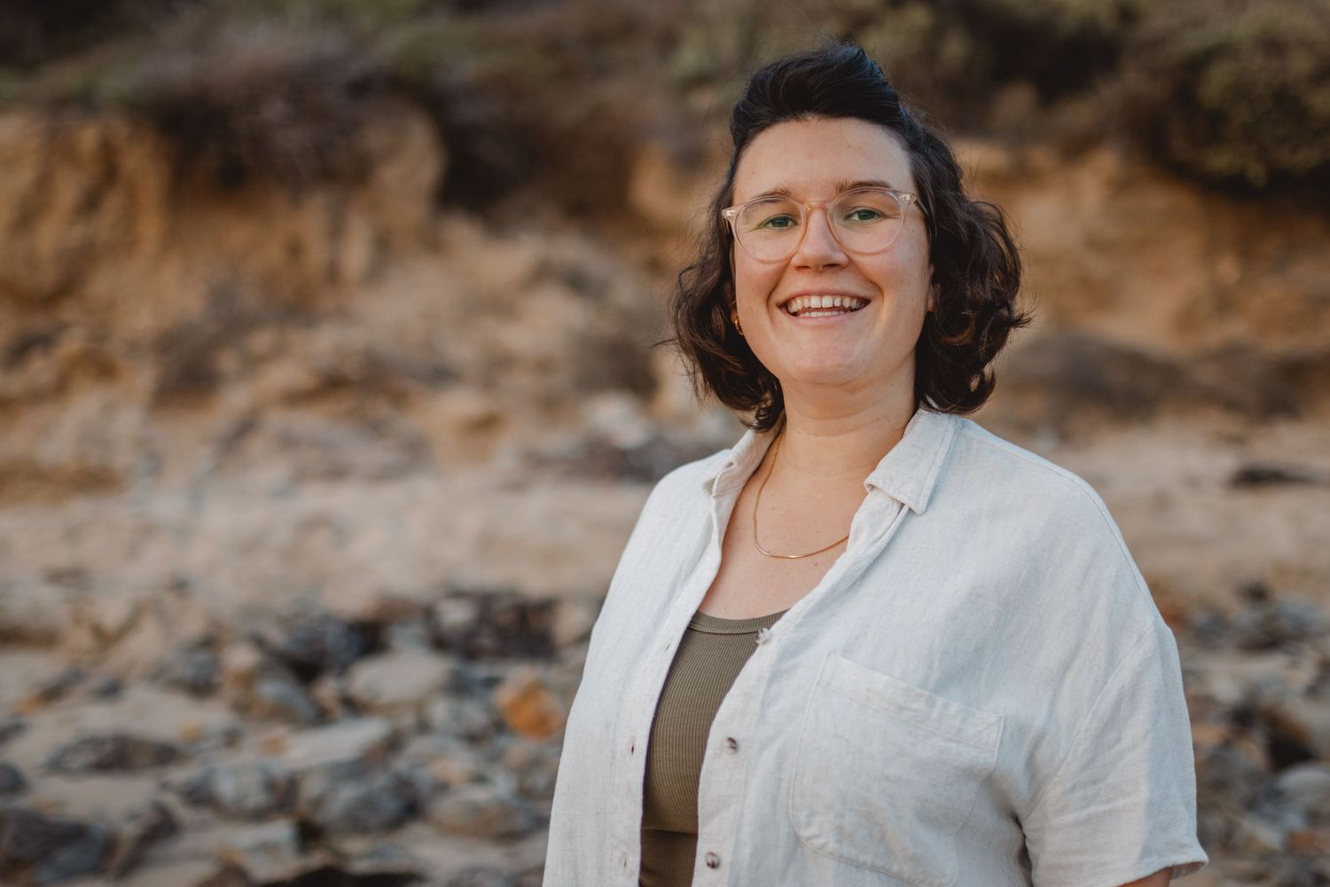A person in a white blazer smiles warmly while standing on a rocky beach with cliffs in the background in Huntington Beach.