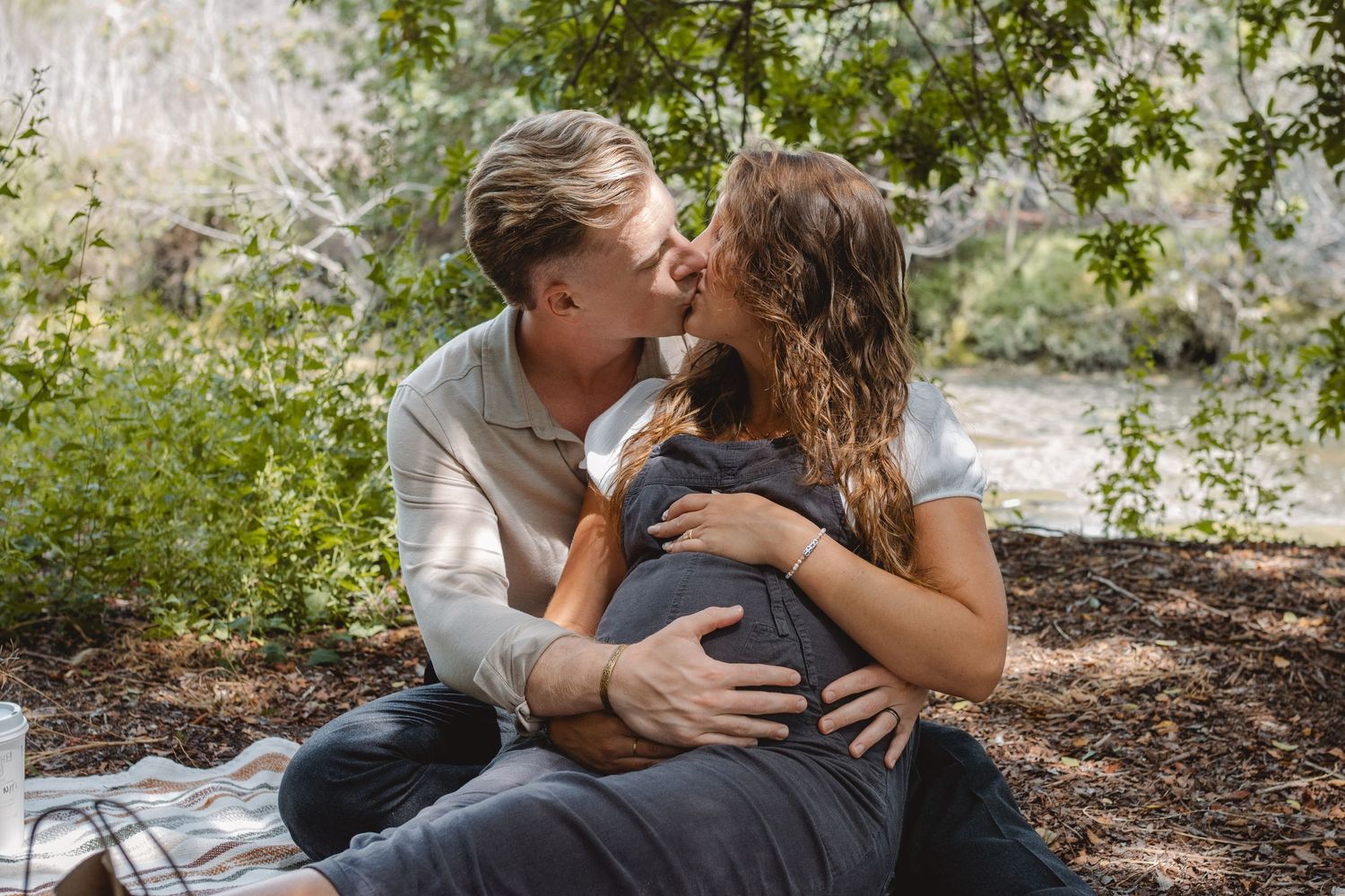 A romantic, pregnant couple sharing intimate moments together under green trees in a wooded outdoor setting in Laguna Beach.