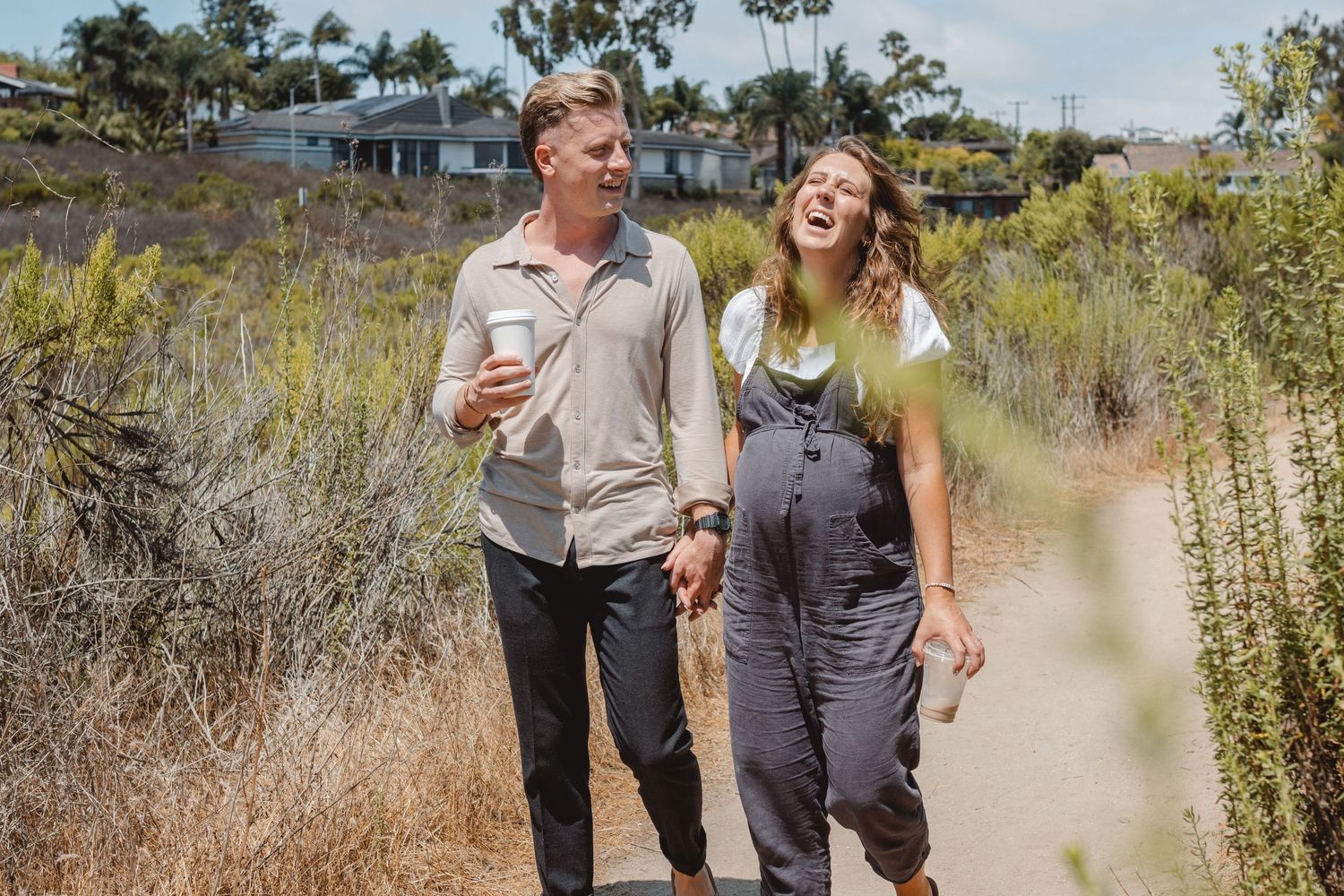 A pregnant couple shares a joyful outdoor moment in Huntington Beach surrounded by desert brush and greenery.