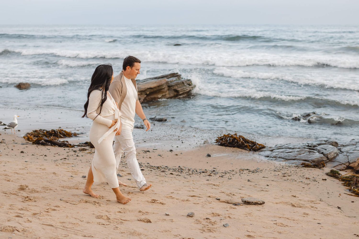 Two people in light colored clothing hold hands while walking along a rocky beach with ocean waves in the background.Newport Pregnancy Shoot
