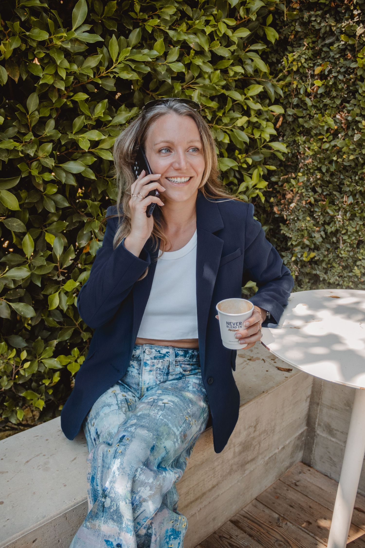Professional wearing navy blazer and tie dye jeans sits outdoors talking on phone while holding coffee cup against leafy backdrop.