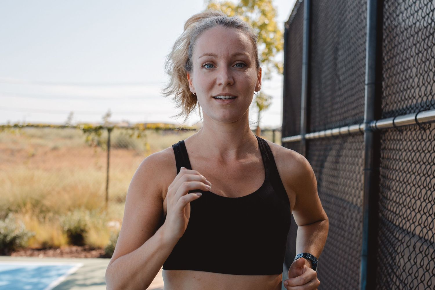 Athletic woman in workout gear smiles while stretching against a chain link fence at an outdoor track in Huntington Beach.