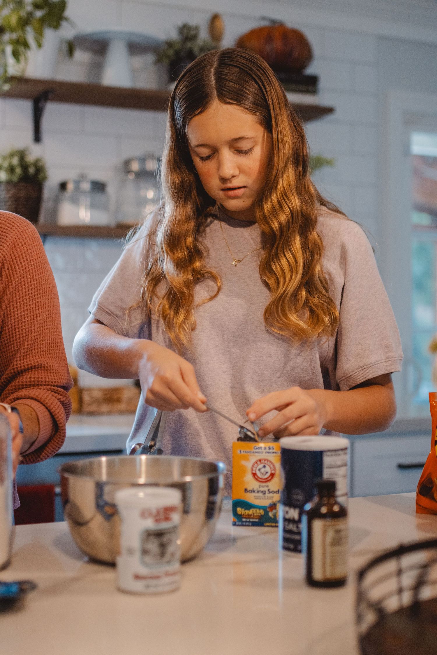 A person with wavy hair in a gray sweater carefully measures ingredients at a kitchen counter while baking in Newport Beach.