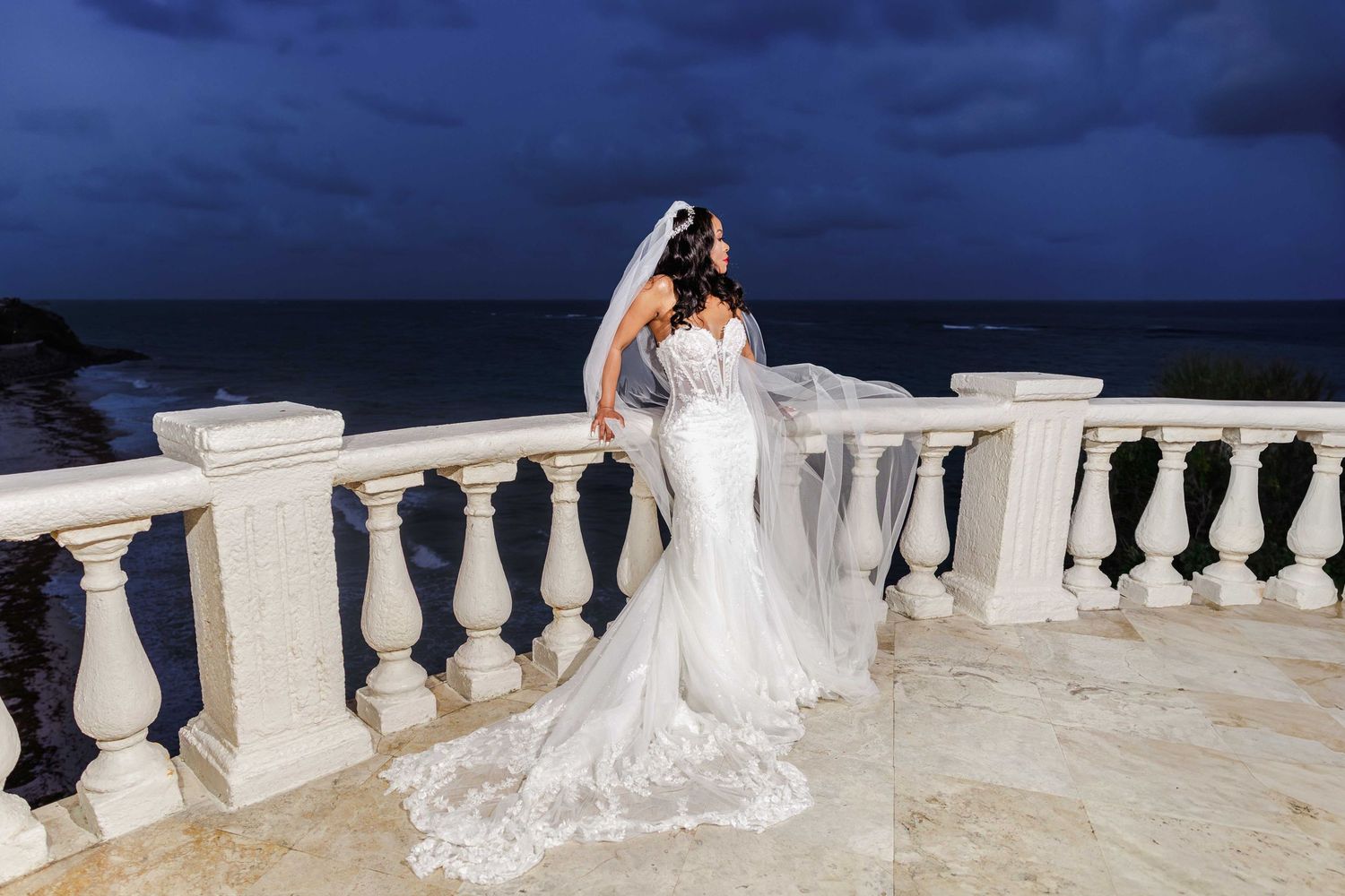 A stunning beachfront wedding scene with white lace gown and veil against a dramatic dark blue ocean backdrop.