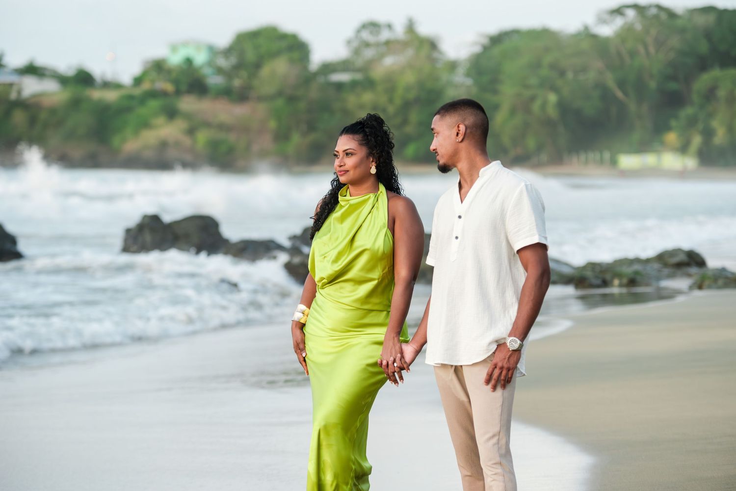 Couple walking hand-in-hand on a tropical beach, woman in green satin dress, man in white shirt, waves crashing on rocks.