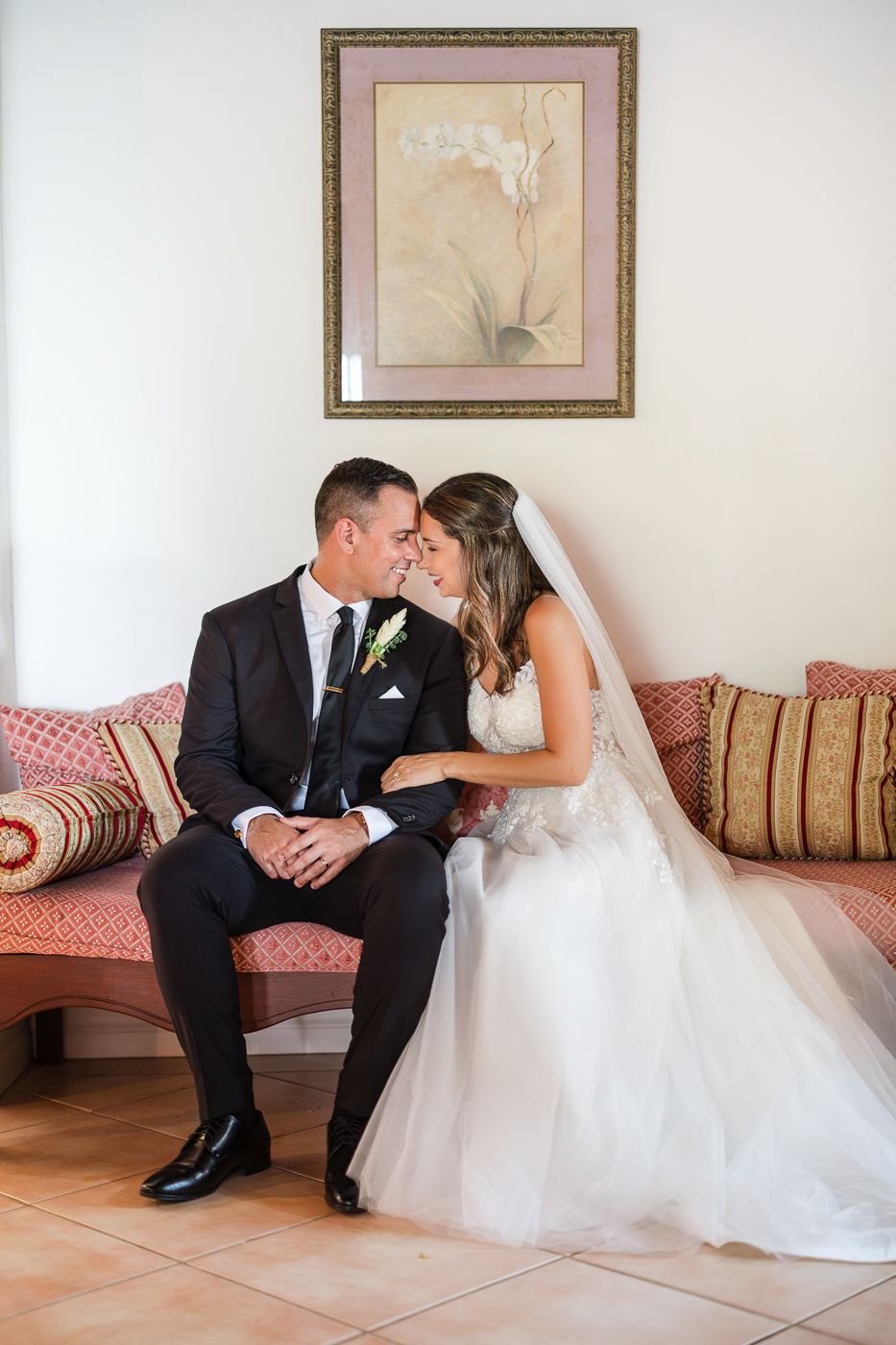 Bride and groom share a tender moment seated on a pink sofa beneath a floral painting on their wedding day.