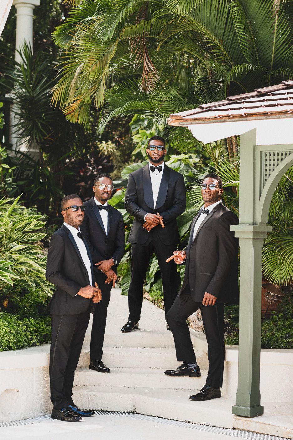 Four groomsmen in black tuxedos and bow ties pose on outdoor steps surrounded by tropical palm trees.