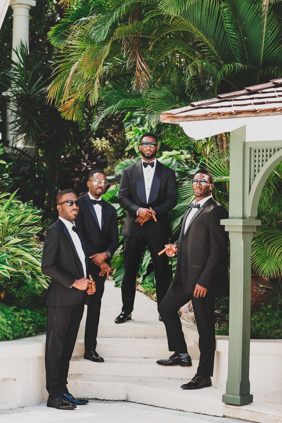 Four groomsmen in black tuxedos and bow ties pose outdoors near a gazebo surrounded by lush tropical greenery.