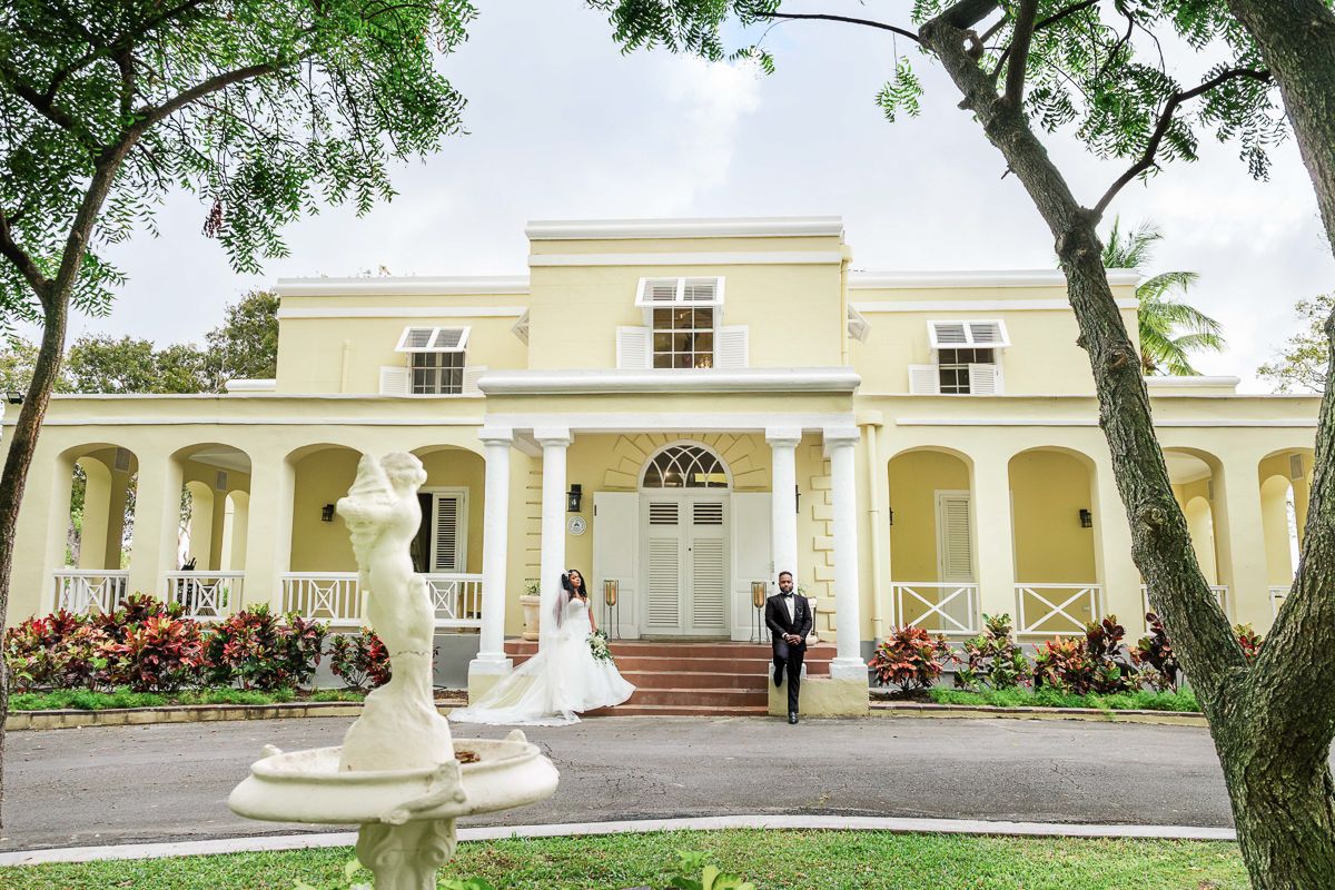 Bride and groom pose outside a elegant yellow colonial mansion with white columns and lush tropical gardens.