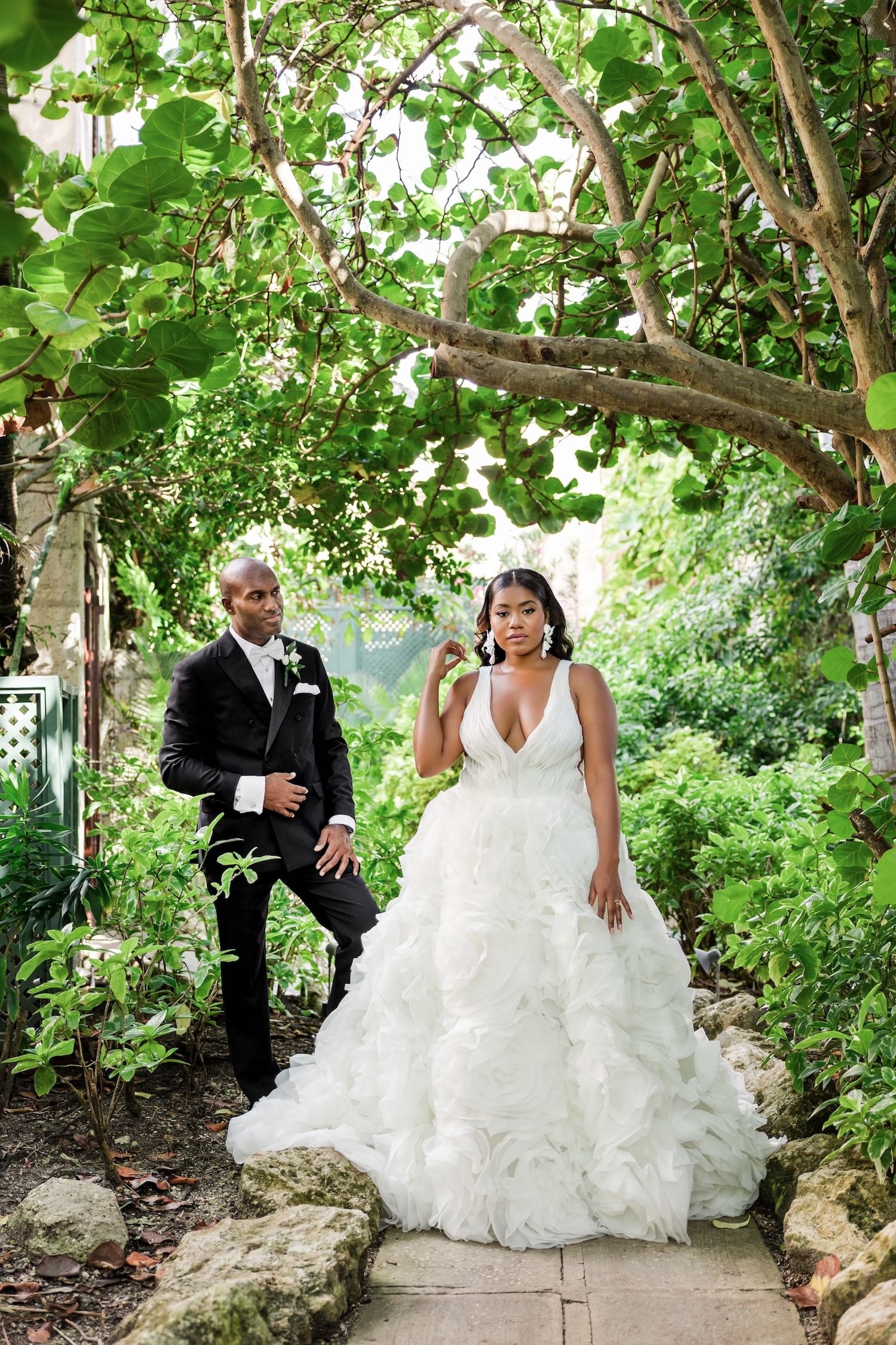 Bride in ruffled ball gown and groom in black tuxedo pose in lush garden archway for elegant wedding portrait.