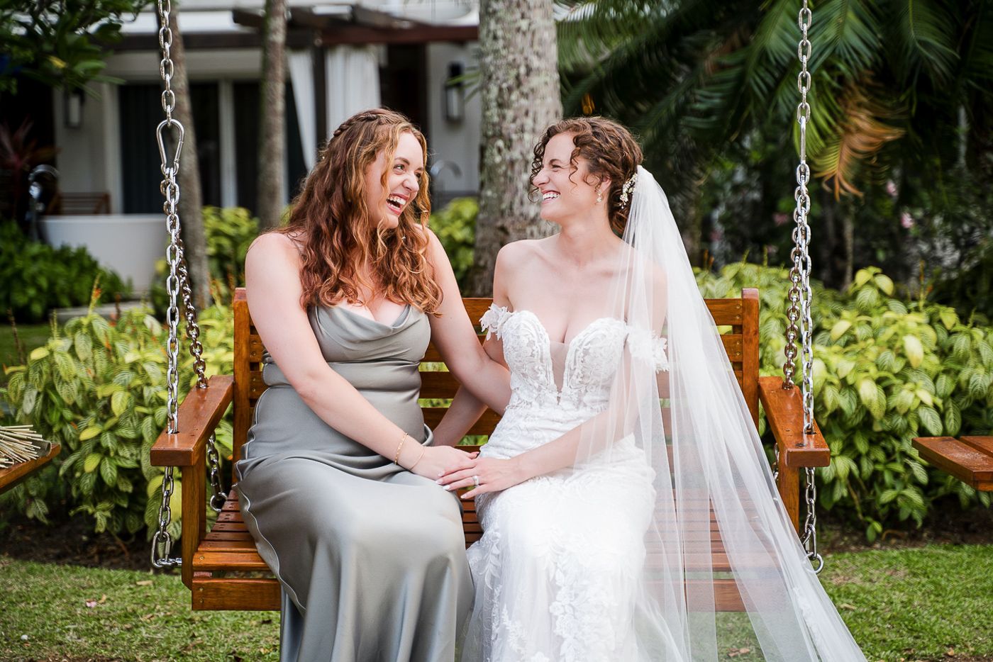 Wedding couple sits intimately on a wooden swing in a lush tropical garden setting.