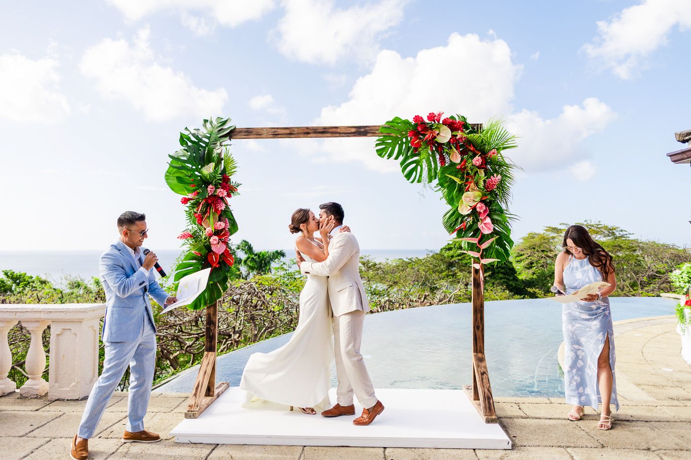 Wedding ceremony under floral arch with ocean view and guests watching.