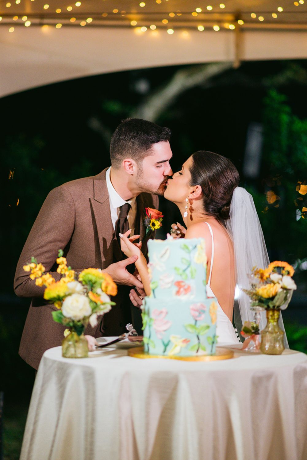 Two people share a romantic kiss at their wedding reception table decorated with yellow flowers and string lights overhead.