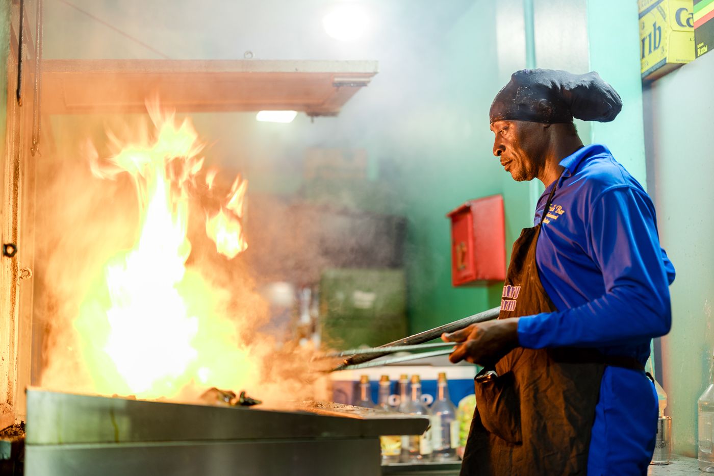 Chef in blue uniform and headband cooking with flames on commercial kitchen grill.