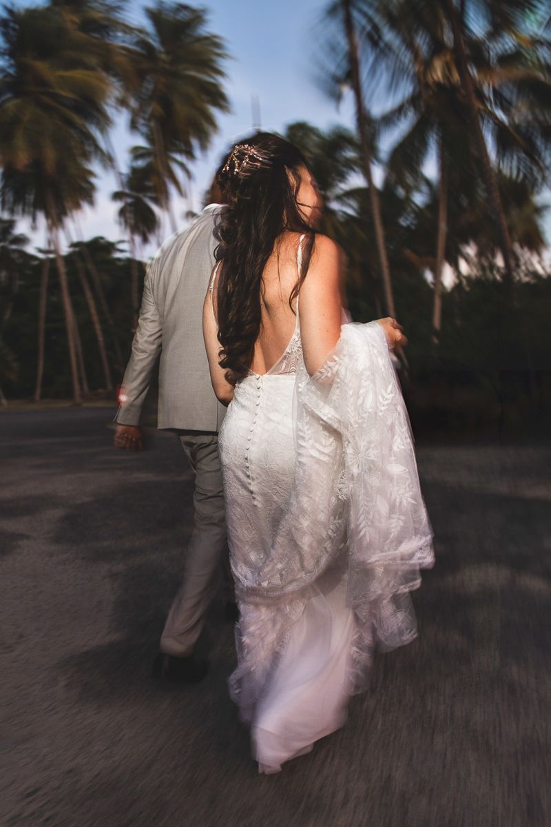 Couple in wedding attire walks along a beach path with palm trees swaying at sunset.