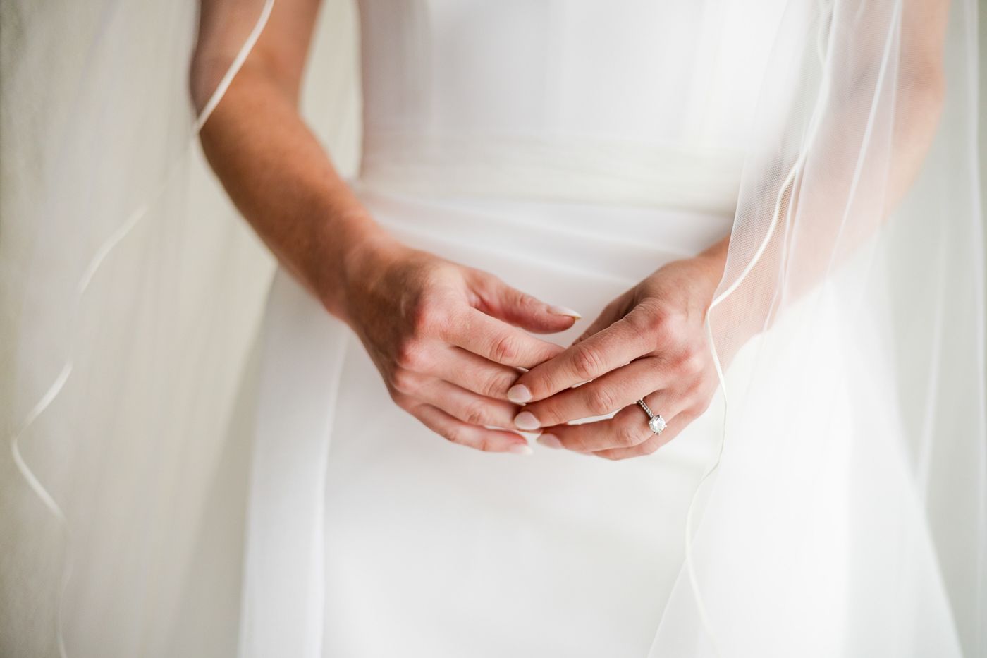 Close-up of a bride in a white wedding dress and veil, hands clasped together showing engagement ring.