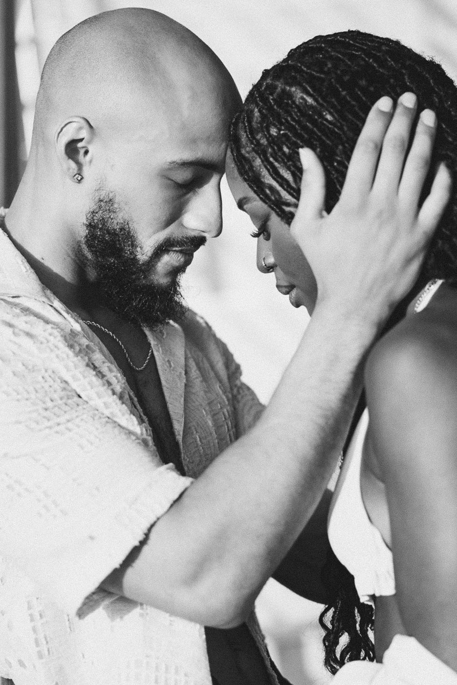 Black and white photo of a bald bearded man tenderly touching foreheads with a woman with braids.