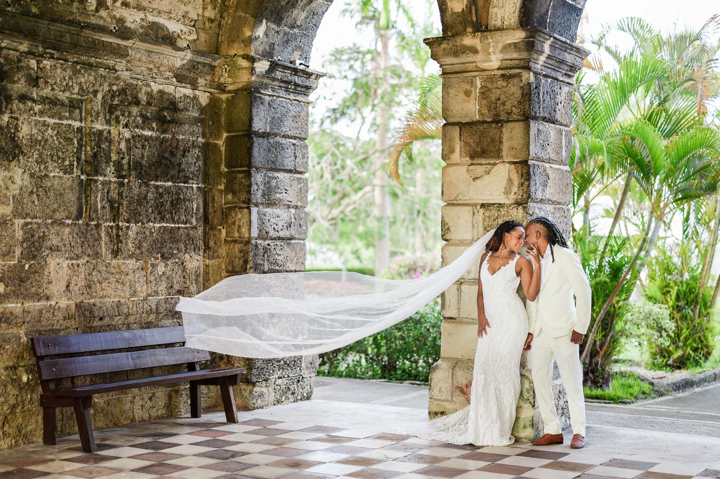 Couple in white wedding attire sharing a romantic moment under stone arches with tropical greenery backdrop.