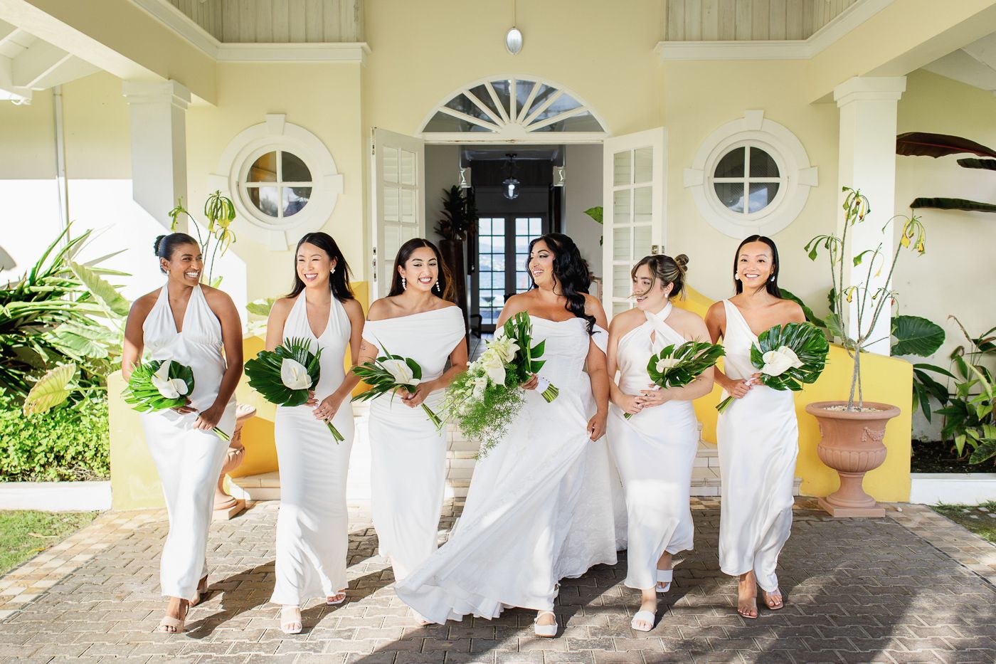 Bride and bridesmaids in white dresses holding tropical bouquets walk together outside a yellow villa.