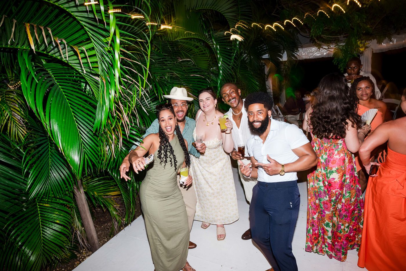 Wedding guests dance and celebrate under tropical palm trees and string lights at an outdoor reception.