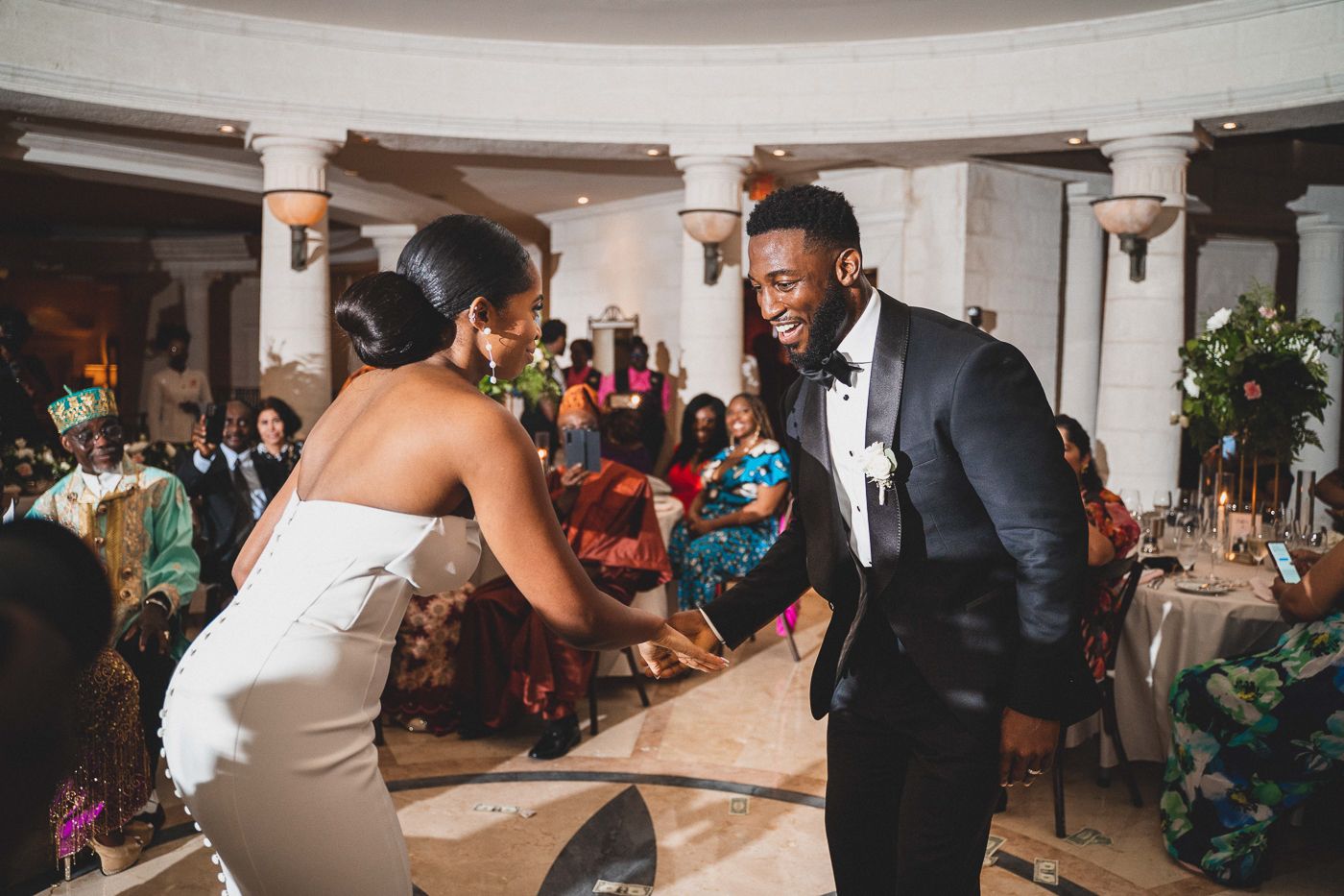 Wedding guests dancing and celebrating in an elegant ballroom with white columns and festive lighting.