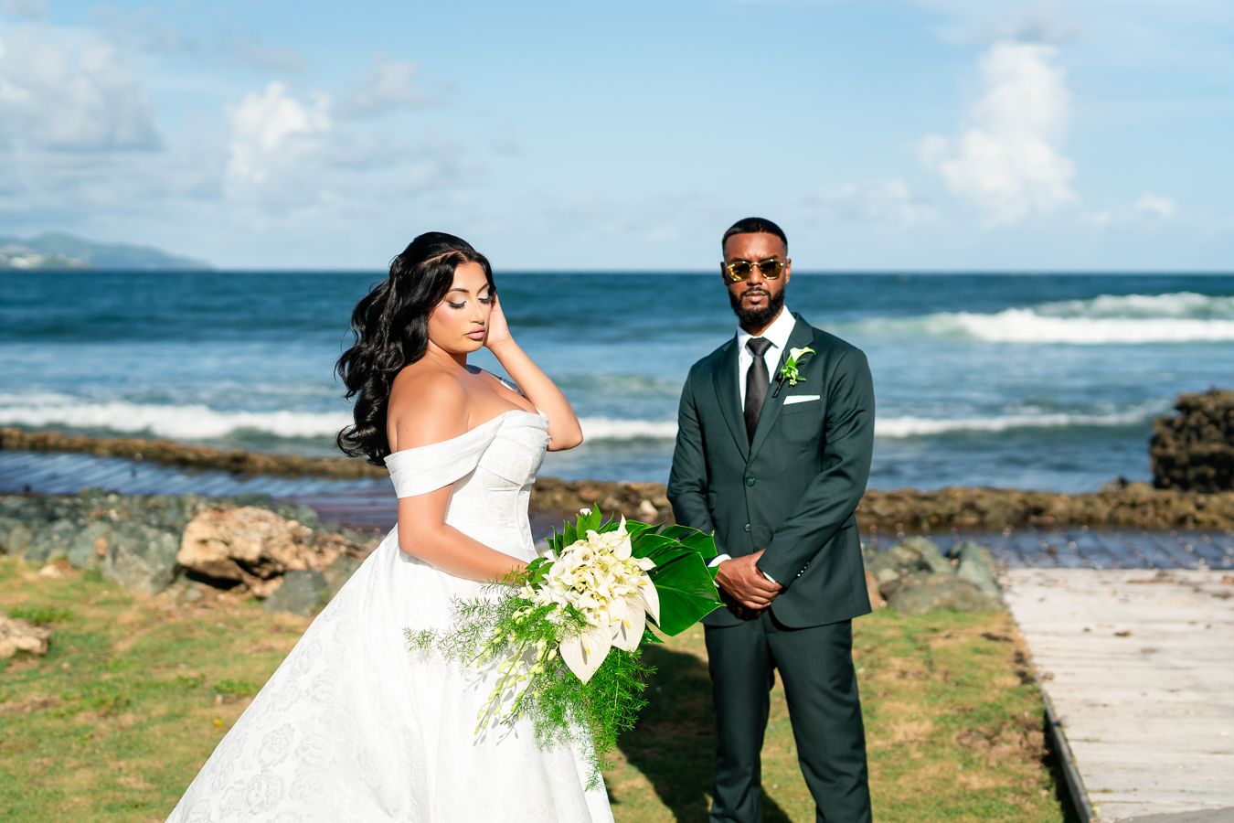A bride in a white dress and groom in dark suit pose beside the ocean on their wedding day.