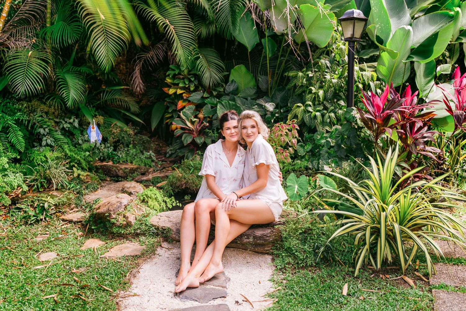 A person sits in a lush tropical garden surrounded by vibrant green plants and pink flowers.