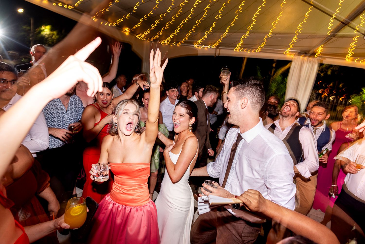 Wedding reception guests dancing in celebration under glowing string lights and decorative white tent canopy.