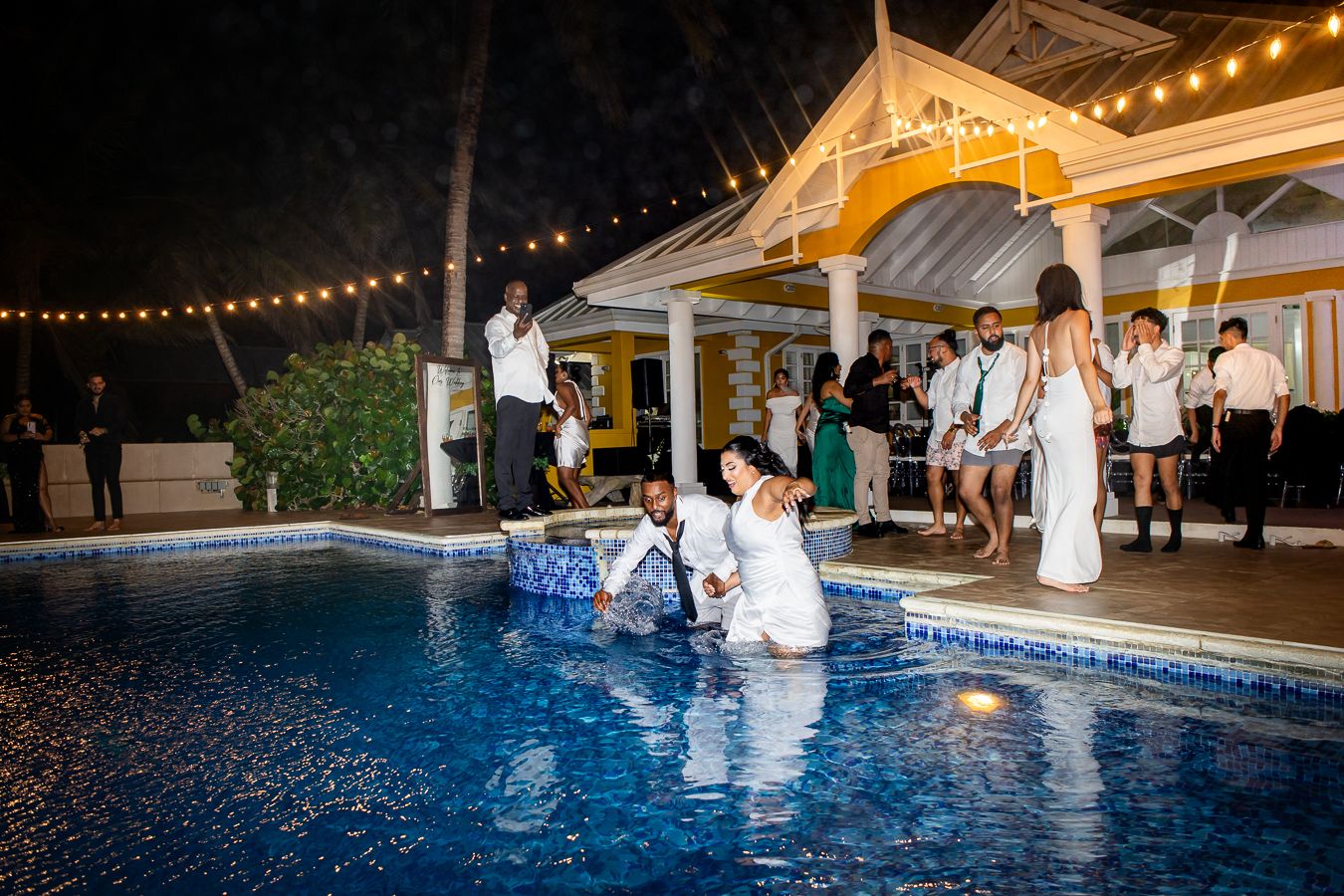 Wedding party jumping into a pool at night with a lit gazebo in the background.