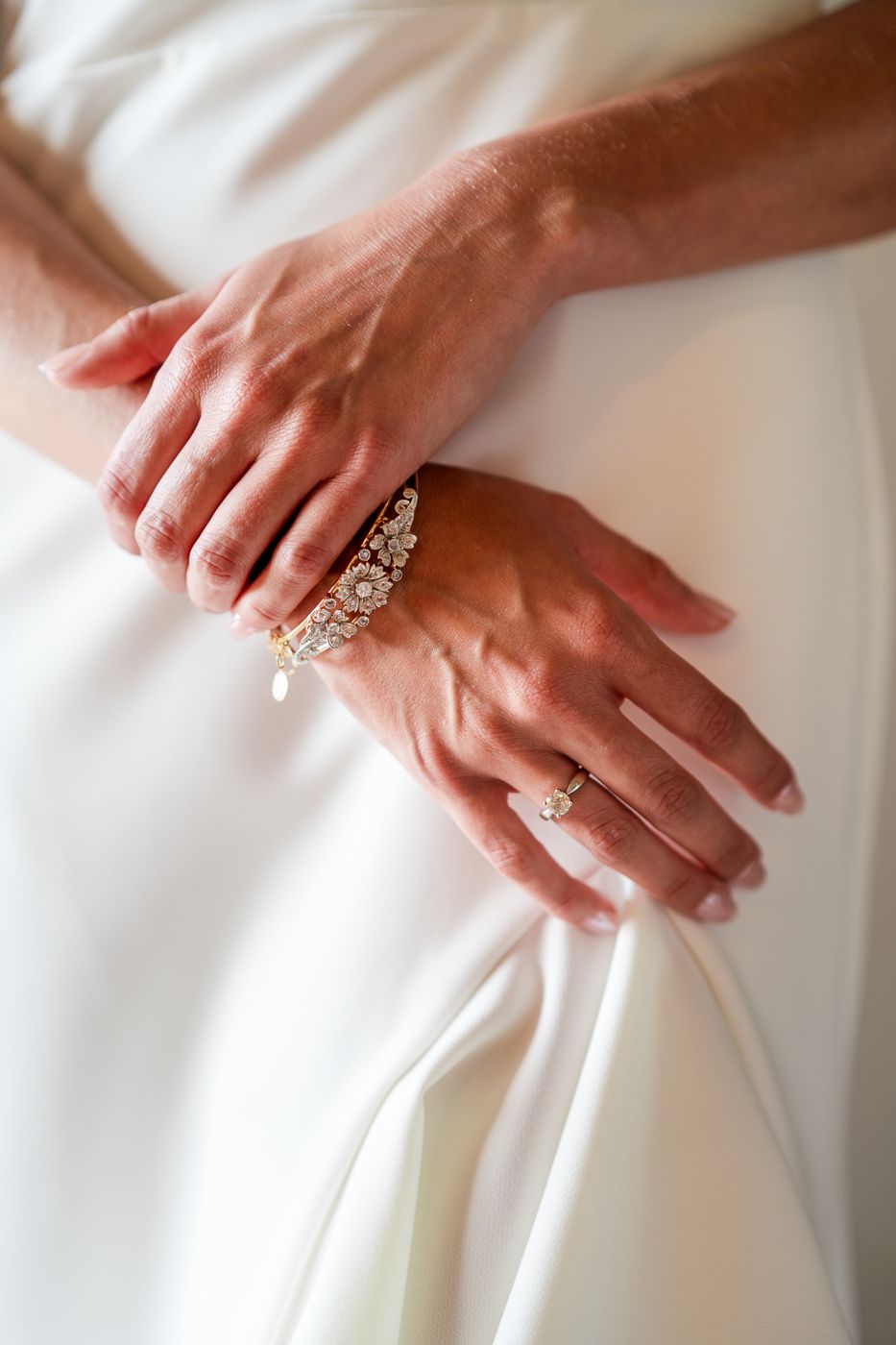 Close-up of hands showing elegant wedding jewelry and bracelet against white dress fabric.