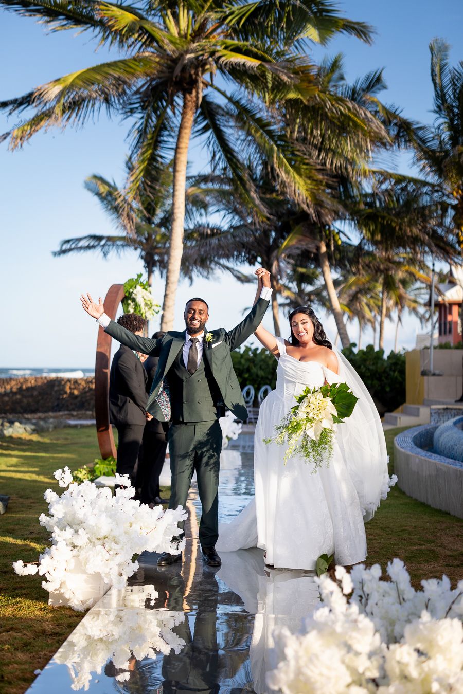 Joyful wedding celebration on reflective white platform with tropical palm trees backdrop.