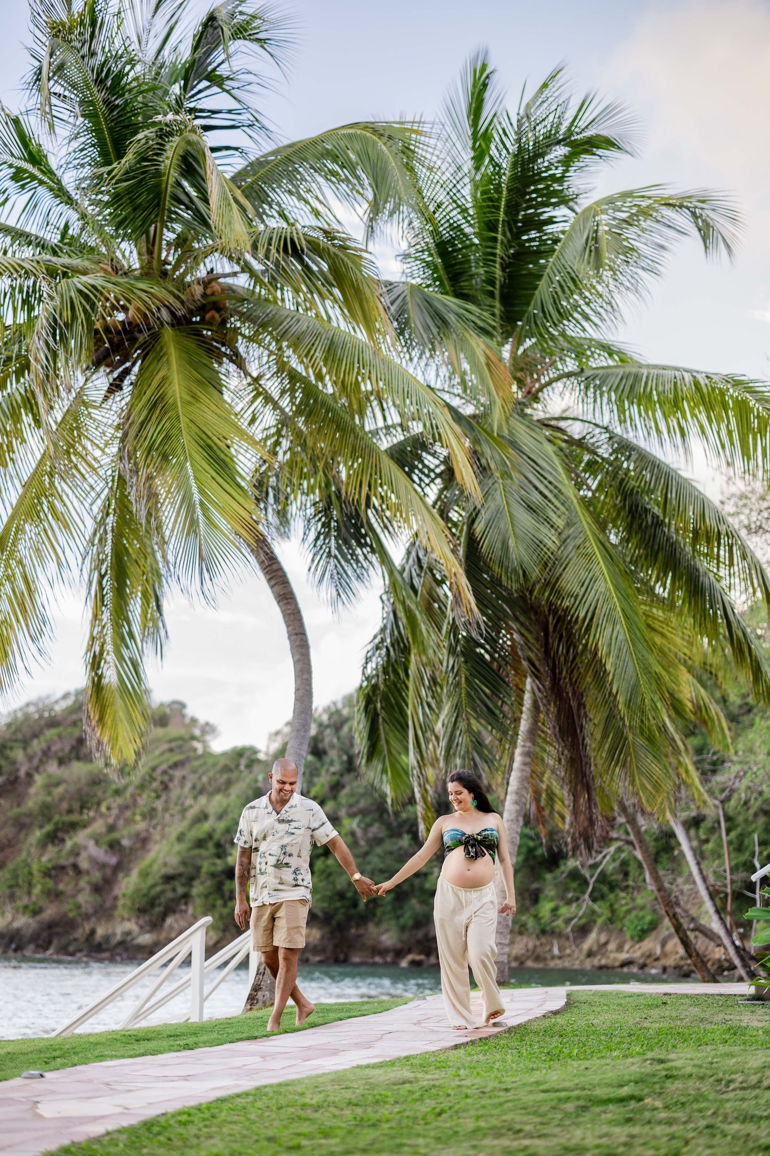Pregnant couple holding hands walking on tropical path with palm trees and ocean views.