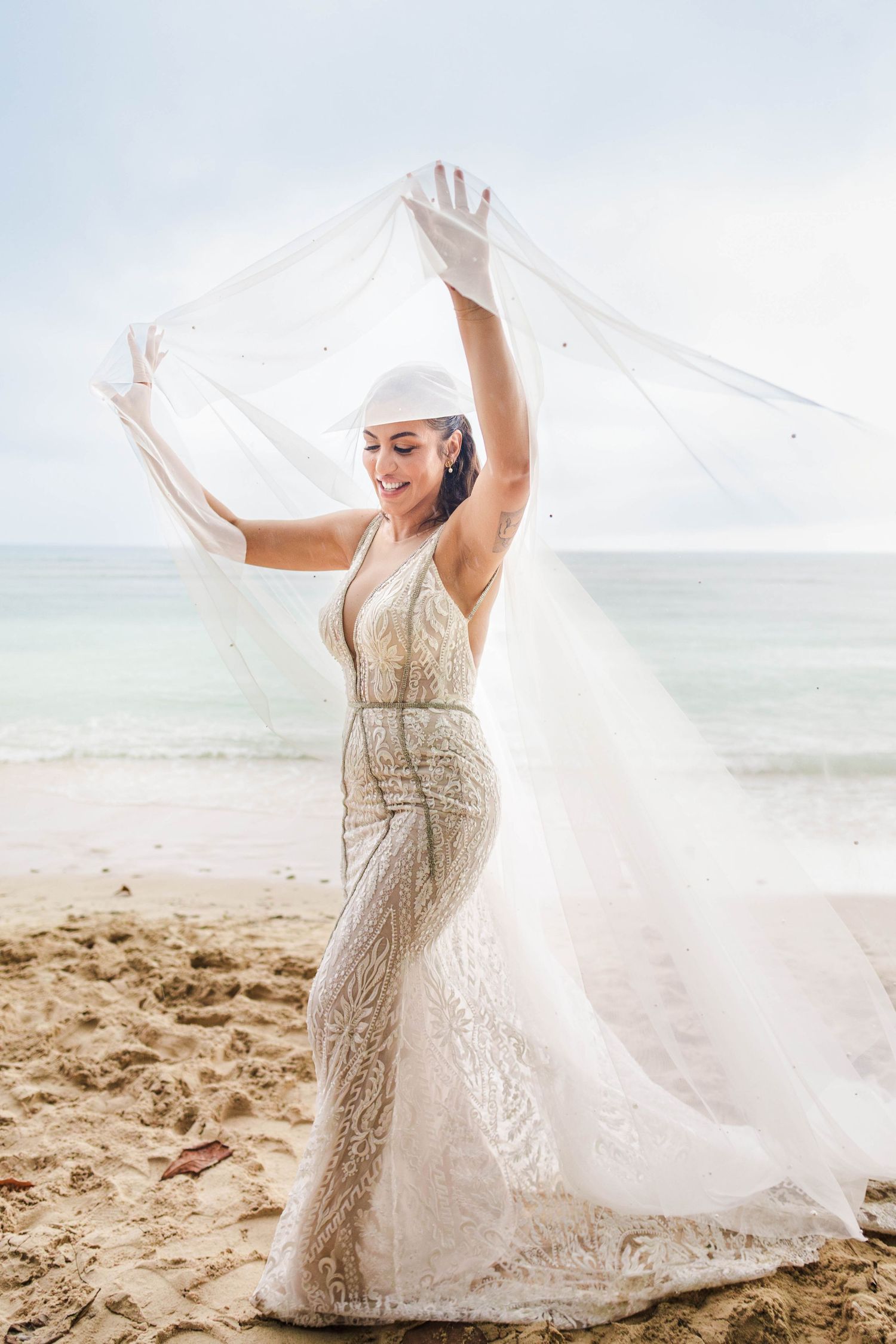Bride in lace gown and white gloves lifts veil on sandy beach with ocean backdrop.