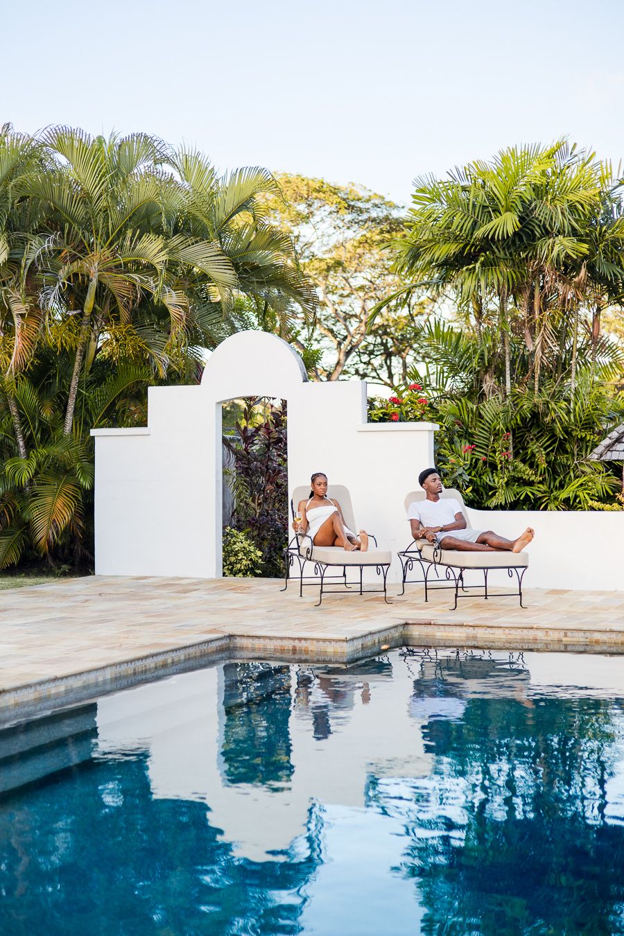 Couple relaxing on lounge chairs by a tropical pool with lush palm trees and white archway in background.