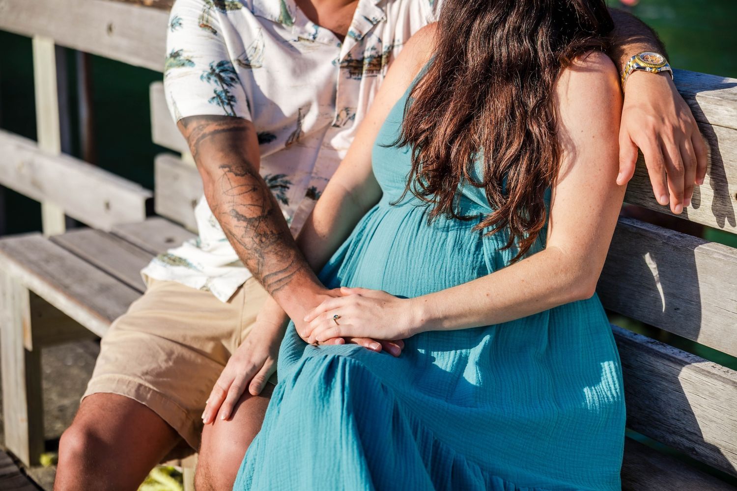 Pregnant woman in teal dress sits with tattooed partner on bench, hands resting on baby bump.