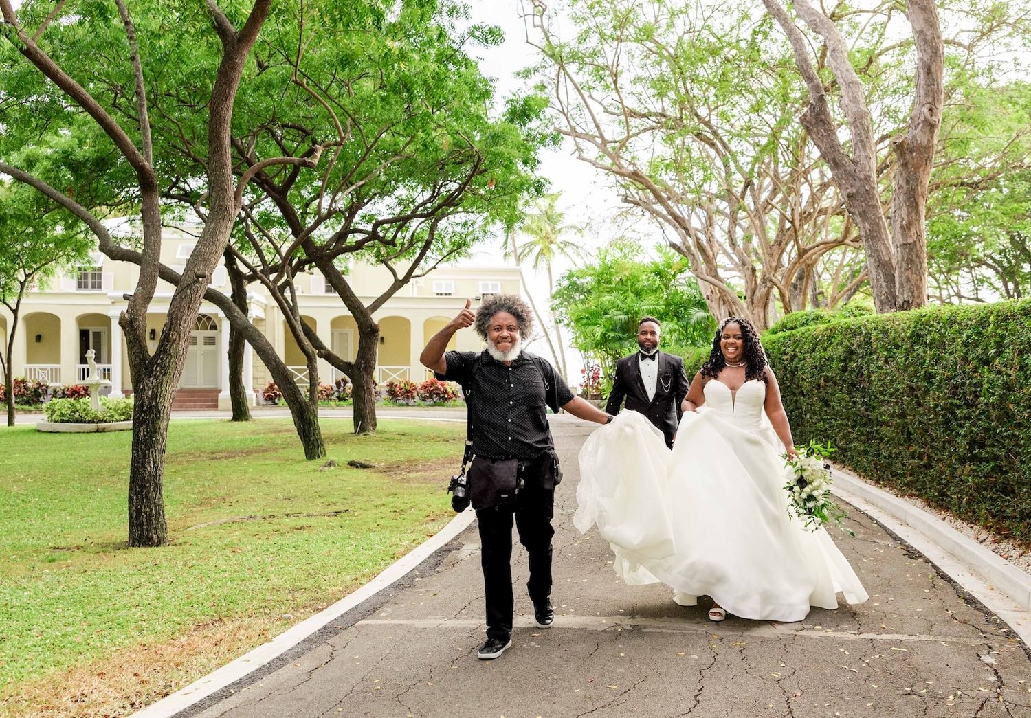 Photographer walks ahead of newlywed couple in white gown and suit on a tree-lined path at outdoor wedding venue.