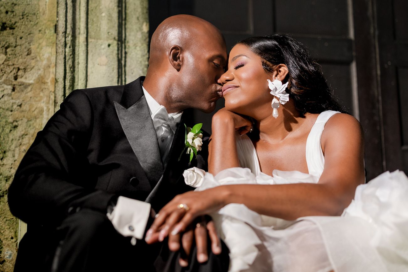 A groom in a black tuxedo gently kisses his bride wearing a white dress and floral hair accessory on their wedding day.