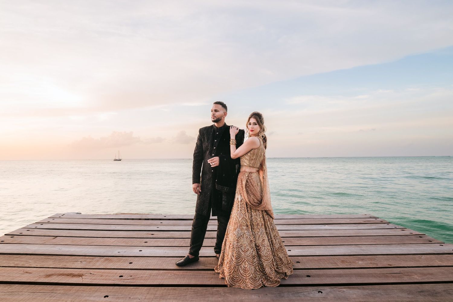 Couple in elegant Indian attire posing on a wooden pier at sunset over calm turquoise ocean waters.